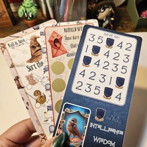 May include: A hand holds several game cards with a fantasy theme. The cards feature illustrations of a sorting hat, an owl, and other magical elements. One card has the text "Roll & Save", and another says "Ronald Weasley". The cards are blue, white, and gold.