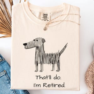 Op de afbeelding: Cr&egrave;mekleurig T-shirt met een zwarte lijntekening van een hond en de tekst "That'll do. I'm Retired." Het shirt is opgevouwen en heeft een Comfort Colors-label. Een zilveren ketting met een hanger ligt erop.