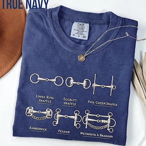 May include: A true navy blue short-sleeve t-shirt featuring a graphic print of six different horse bits with labels: Loose Ring Snaffle, Eggrutt Snaffle, Full Cheen Snaffle, Kimberwick, Pelham, and Wetmouth & Bradoon. The shirt has a crew neckline.