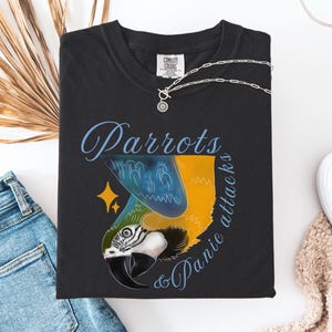 May include: Black t-shirt featuring a parrot design in blue, yellow, and white, with the text "Parrots & Panic Attacks" in blue script. A silver necklace with a sun pendant is visible.