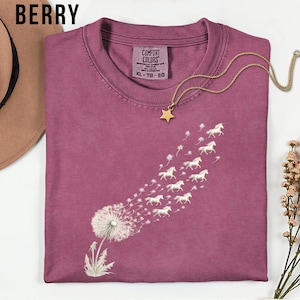 May include: Berry-coloured t-shirt featuring a dandelion design, with white horses replacing the seeds. A gold star pendant necklace is placed on the shirt. The shirt has a crew neck and the tag reads "COMFORT COLORS" and "XL - TG - EG".