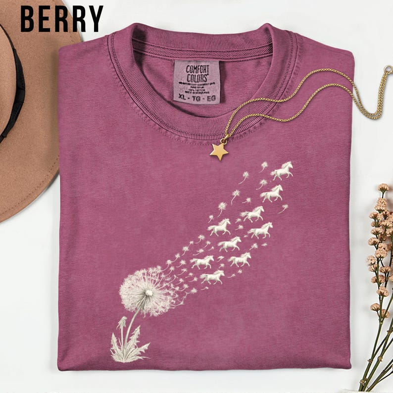 May include: Berry-coloured t-shirt featuring a dandelion design, with white horses replacing the seeds. A gold star pendant necklace is placed on the shirt. The shirt has a crew neck and the tag reads "COMFORT COLORS" and "XL - TG - EG".