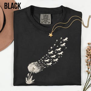 May include: Black crew neck t-shirt featuring a dandelion design. The dandelion seeds morph into white horses in flight. A gold star pendant necklace is on the shirt. The shirt has a Comfort Colors tag and the word "BLACK" is at the top.