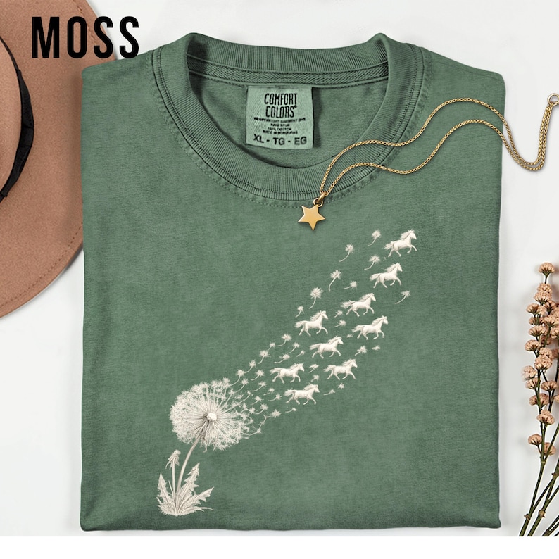 May include: A moss green t-shirt featuring a dandelion design, with the seeds morphing into white horses. The shirt has a crew neck and short sleeves. A gold star pendant necklace is on the shirt. The tag reads "Comfort Colors" and "XL - TG - EG".