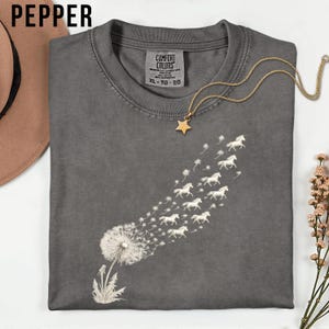 May include: A grey t-shirt featuring a dandelion design, with the seeds morphing into white horses. A gold star pendant necklace and a brown hat are also visible. The shirt is labelled "Comfort Colors" and is size XL-TG-EG.