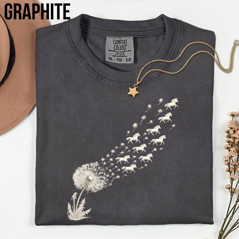 May include: Graphite-coloured t-shirt featuring a dandelion design. The dandelion seeds morph into white horses, forming a whimsical graphic. A gold star pendant necklace is on the shirt. The shirt is labelled "Comfort Colors" and is size XL-TG-EG.