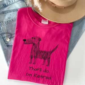 Op de afbeelding: Een felroze t-shirt met een zwarte hondenillustratie en de tekst "That'll do. I'm Retired." Het shirt ligt op een blauwe spijkerbroek, met een beige hoed op de achtergrond.