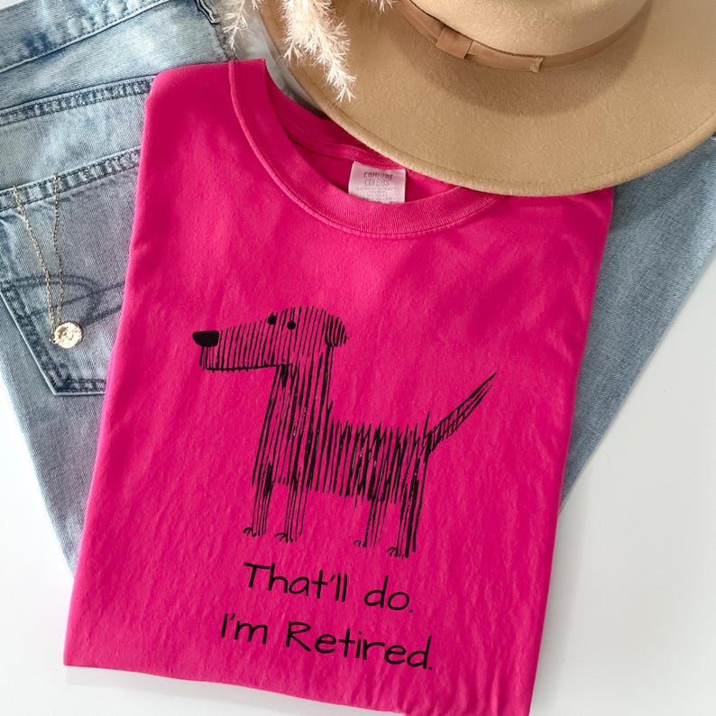 Op de afbeelding: Een felroze t-shirt met een zwarte hondenillustratie en de tekst "That'll do. I'm Retired." Het shirt ligt op een blauwe spijkerbroek, met een beige hoed op de achtergrond.