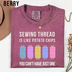 May include: Berry-colored t-shirt with the text "SEWING THREAD IS LIKE POTATO CHIPS YOU CAN'T HAVE JUST ONE." The shirt features colorful spools of thread. A gold star pendant necklace is draped across the shirt.