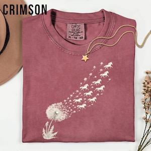 May include: Crimson t-shirt with a dandelion design and white horses blowing away. The shirt has a crew neck and short sleeves. A gold star pendant necklace is draped across the shirt. The tag reads "Comfort Colors" and "XL - TG - EG".