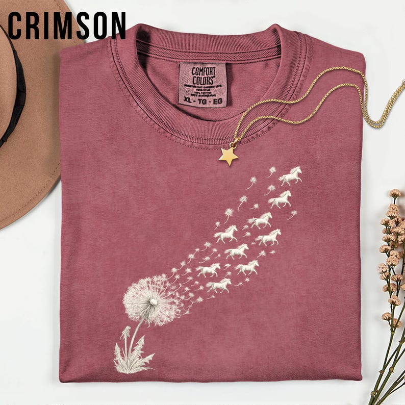 May include: Crimson t-shirt with a dandelion design and white horses blowing away. The shirt has a crew neck and short sleeves. A gold star pendant necklace is draped across the shirt. The tag reads "Comfort Colors" and "XL - TG - EG".