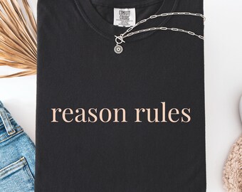 May include: A black short-sleeved t-shirt with the text "reason rules" printed in a light peach color across the chest. The shirt features a crew neckline and a Comfort Colors brand label at the inner collar. A silver-toned chain necklace with a circular pendant rests on the neckline of the shirt.