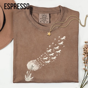 May include: Espresso-coloured t-shirt featuring a dandelion design. The dandelion seeds morph into a flock of white horses. A gold star pendant necklace rests on the shirt. The shirt is labelled XL-TG-EG.