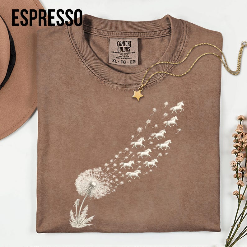 May include: Espresso-coloured t-shirt featuring a dandelion design. The dandelion seeds morph into a flock of white horses. A gold star pendant necklace rests on the shirt. The shirt is labelled XL-TG-EG.