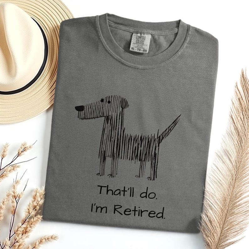 Op de afbeelding: Grijs t-shirt met een zwarte hondenillustratie en de tekst "That'll do. I'm Retired." Het shirt heeft een ronde hals en korte mouwen. Een strohoed en een decoratieve plant staan op de achtergrond.
