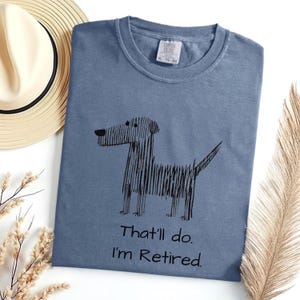 Op de afbeelding: Een stoffig blauw t-shirt met een zwarte schets van een hond en de tekst "That'll do. I'm Retired." Een strohoed en een gedroogde plant liggen naast het t-shirt.