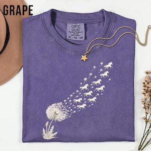 May include: A grape-coloured t-shirt featuring a dandelion design, with the seeds morphing into white horses. The shirt has a crew neck and a "Comfort Colors" tag. A gold necklace with a star pendant is draped on the shirt.