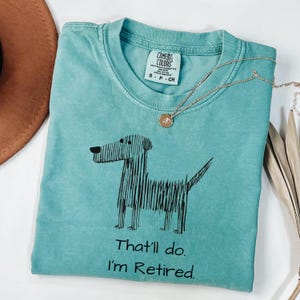 Op de afbeelding: Een turquoise Comfort Colors t-shirt met een zwarte hondenafbeelding en de tekst "That'll do. I'm Retired." Een gouden ketting met een hanger ligt op het shirt. Het shirt is maat S.