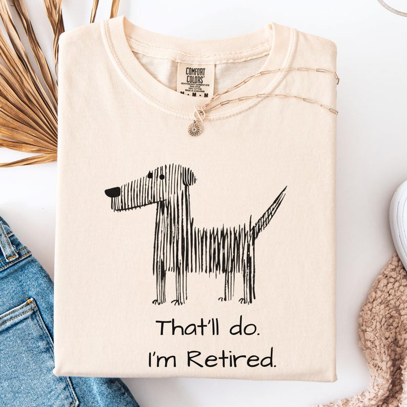 Op de afbeelding: Cr&egrave;mekleurig T-shirt met een zwarte lijntekening van een hond en de tekst "That'll do. I'm Retired." Het shirt is opgevouwen en heeft een Comfort Colors-label. Een zilveren ketting met een hanger ligt erop.