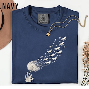 May include: A navy blue t-shirt featuring a dandelion design with white horse silhouettes replacing the seeds. A gold star pendant necklace is placed on the shirt. The shirt has a Comfort Colors tag. The word "NAVY" is printed in the upper left corner.