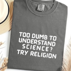 May include: A gray t-shirt with the text "TOO DUMB TO UNDERSTAND SCIENCE? TRY RELIGION" in white block letters. The shirt is folded and has a tag in the upper center. A straw hat and decorative dried plants are in the background.