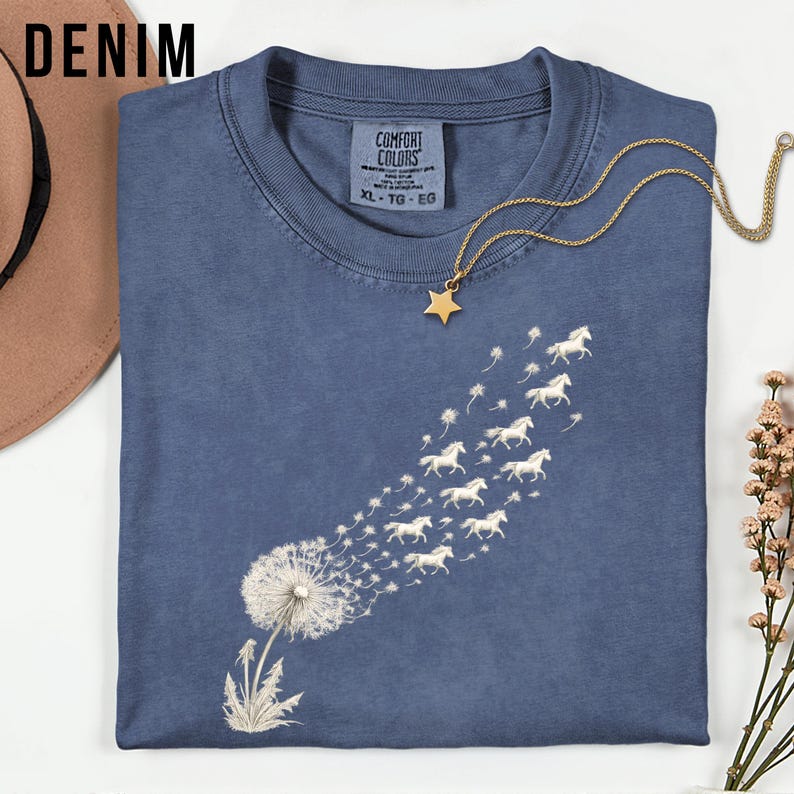 May include: A denim blue t-shirt featuring a dandelion design, with the seeds morphing into white horses. The shirt has a crew neck and short sleeves. A gold necklace with a star pendant is on the shirt. The word "DENIM" is in the upper left corner.