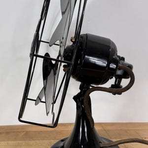 Pu&ograve; includere: Un antico ventilatore elettrico nero con una gabbia e tre pale. Il ventilatore ha una finitura lucida e un cavo di alimentazione marrone. Il ventilatore &egrave; posto su una superficie di legno.