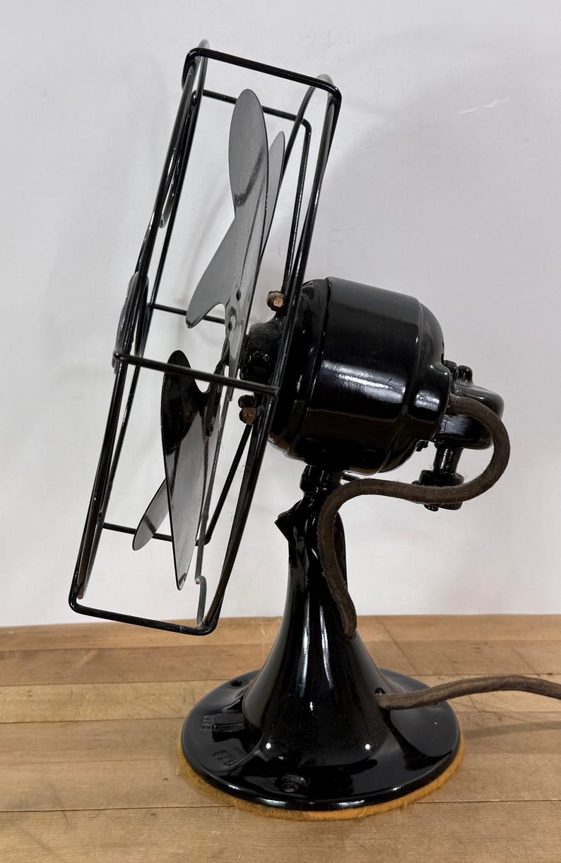 Pu&ograve; includere: Un antico ventilatore elettrico nero con una gabbia e tre pale. Il ventilatore ha una finitura lucida e un cavo di alimentazione marrone. Il ventilatore &egrave; posto su una superficie di legno.