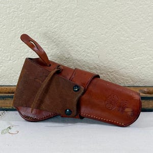 May include: A brown leather holster with a loop and a flap. The holster has a cylindrical shape with a stitched design and a dark brown suede flap. The leather is a rich, reddish-brown color. The holster appears to be vintage.