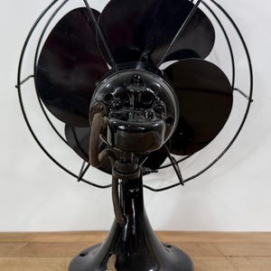 Pu&ograve; includere: Ventilatore elettrico nero vintage con tre pale, una gabbia protettiva in filo metallico e finitura lucida. Il ventilatore ha una base arrotondata e un cavo. Il ventilatore &egrave; un oggetto d'antiquariato.