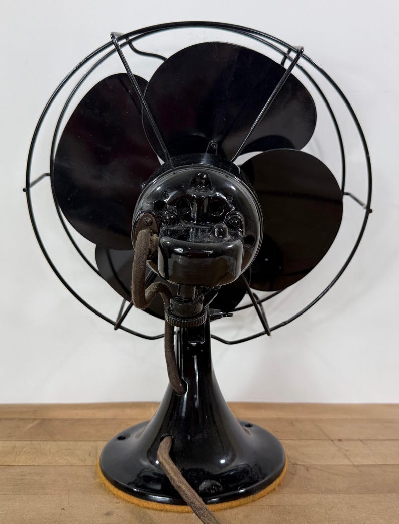Pu&ograve; includere: Ventilatore elettrico nero vintage con tre pale, una gabbia protettiva in filo metallico e finitura lucida. Il ventilatore ha una base arrotondata e un cavo. Il ventilatore &egrave; un oggetto d'antiquariato.