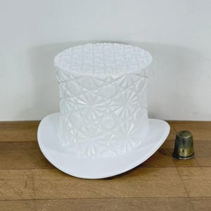 May include: A white milk glass top hat with a geometric pressed pattern featuring starbursts and diamonds across the cylindrical crown and flat top. The brim is smooth and curved, and a small metallic thimble sits beside the glass object.