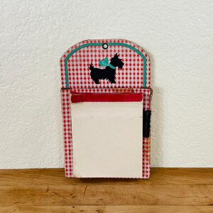 May include: A vintage-style notepad holder with a red and white gingham pattern. The top features a black silhouette of a dog with a teal bow. The notepad has a red bar at the top and a blank white pad for notes.