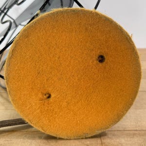 Pu&ograve; includere: Primo piano della base di un ventilatore vintage in metallo nero. La base &egrave; un cuscinetto rotondo in feltro color senape con due piccoli fori. Una gabbia e le pale in metallo nero sono visibili sullo sfondo.