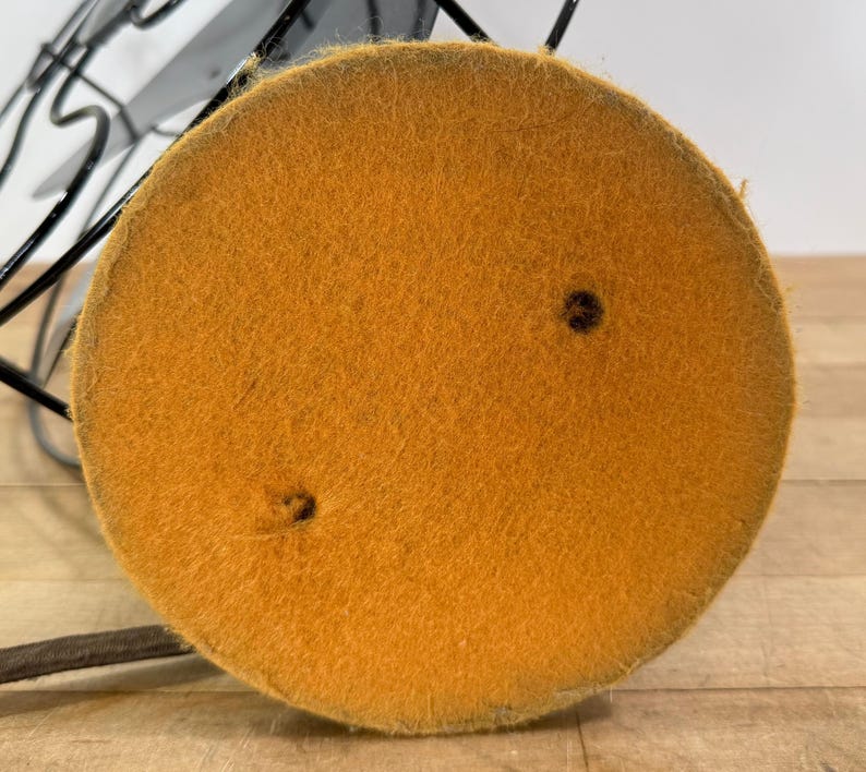 Pu&ograve; includere: Primo piano della base di un ventilatore vintage in metallo nero. La base &egrave; un cuscinetto rotondo in feltro color senape con due piccoli fori. Una gabbia e le pale in metallo nero sono visibili sullo sfondo.