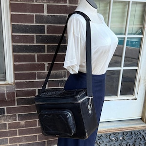 May include: Black rectangular cooler bag with a shoulder strap. The bag has a front pocket and a zipper closure. The bag is worn over the shoulder of a mannequin wearing a white shirt and navy blue skirt.