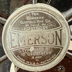 Pu&ograve; includere: Primo piano dell'etichetta di un motore di ventilatore Emerson vintage. L'etichetta circolare presenta il marchio "EMERSON" a caratteri grandi, insieme al testo che indica il produttore, la posizione e le specifiche tecniche. L'etichetta &egrave; metallica con un aspetto usurato.