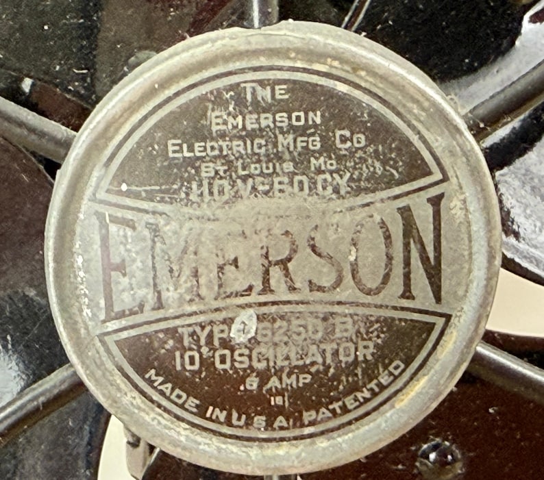 Pu&ograve; includere: Primo piano dell'etichetta di un motore di ventilatore Emerson vintage. L'etichetta circolare presenta il marchio "EMERSON" a caratteri grandi, insieme al testo che indica il produttore, la posizione e le specifiche tecniche. L'etichetta &egrave; metallica con un aspetto usurato.