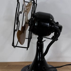 May include: An antique black electric fan with a cage and three blades. The fan has a glossy finish and a round base. The blades are a light brown colour. The fan is on a wooden surface.