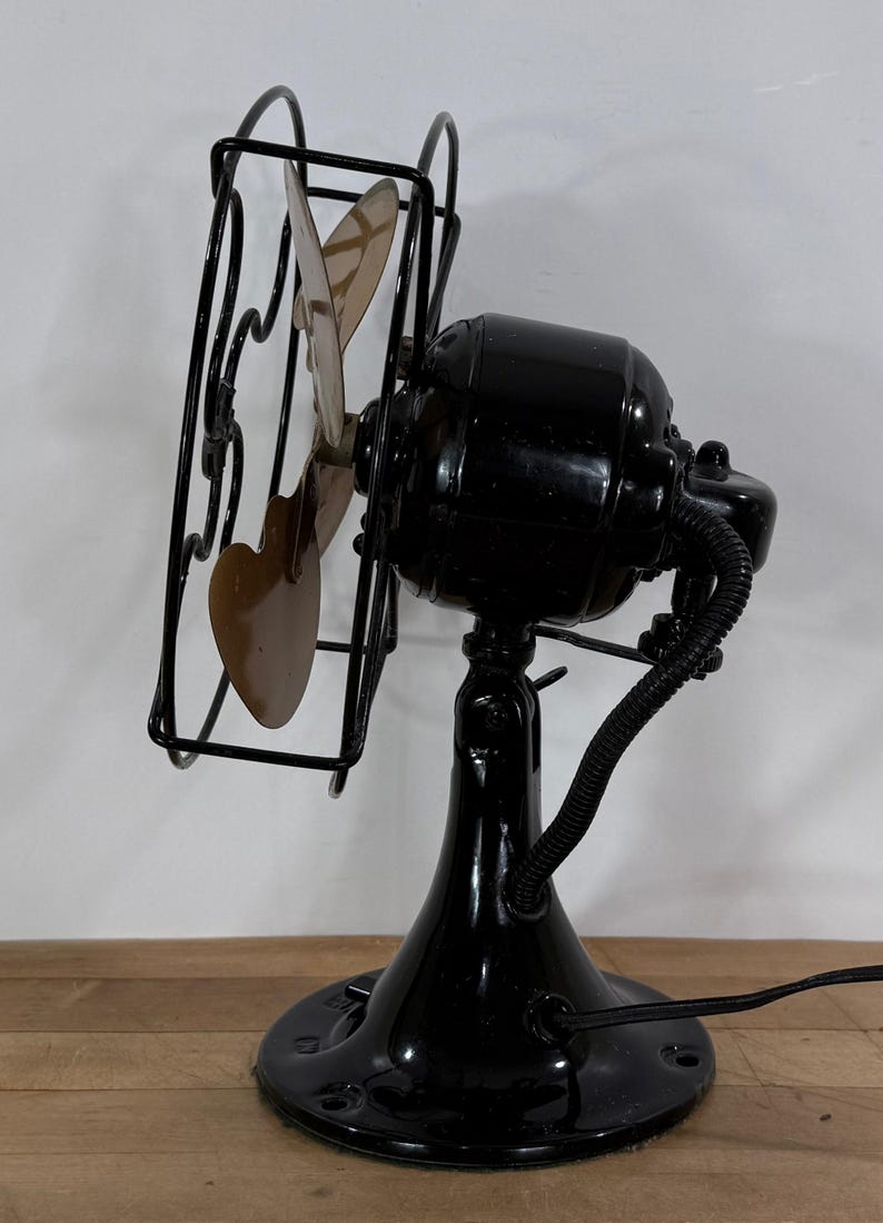 May include: An antique black electric fan with a cage and three blades. The fan has a glossy finish and a round base. The blades are a light brown colour. The fan is on a wooden surface.