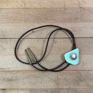 May include: A bolo tie featuring a braided dark brown leather cord with antique brass metal tips. The central slide is a triangular piece of light mint green material centered with a circular silver-toned coin depicting a profile portrait.