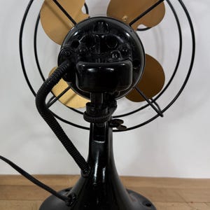 May include: A vintage black electric fan with three gold-coloured blades, enclosed in a black wire cage. The fan has a black base and motor housing, with a flexible cord extending from the back. The fan is a classic design.