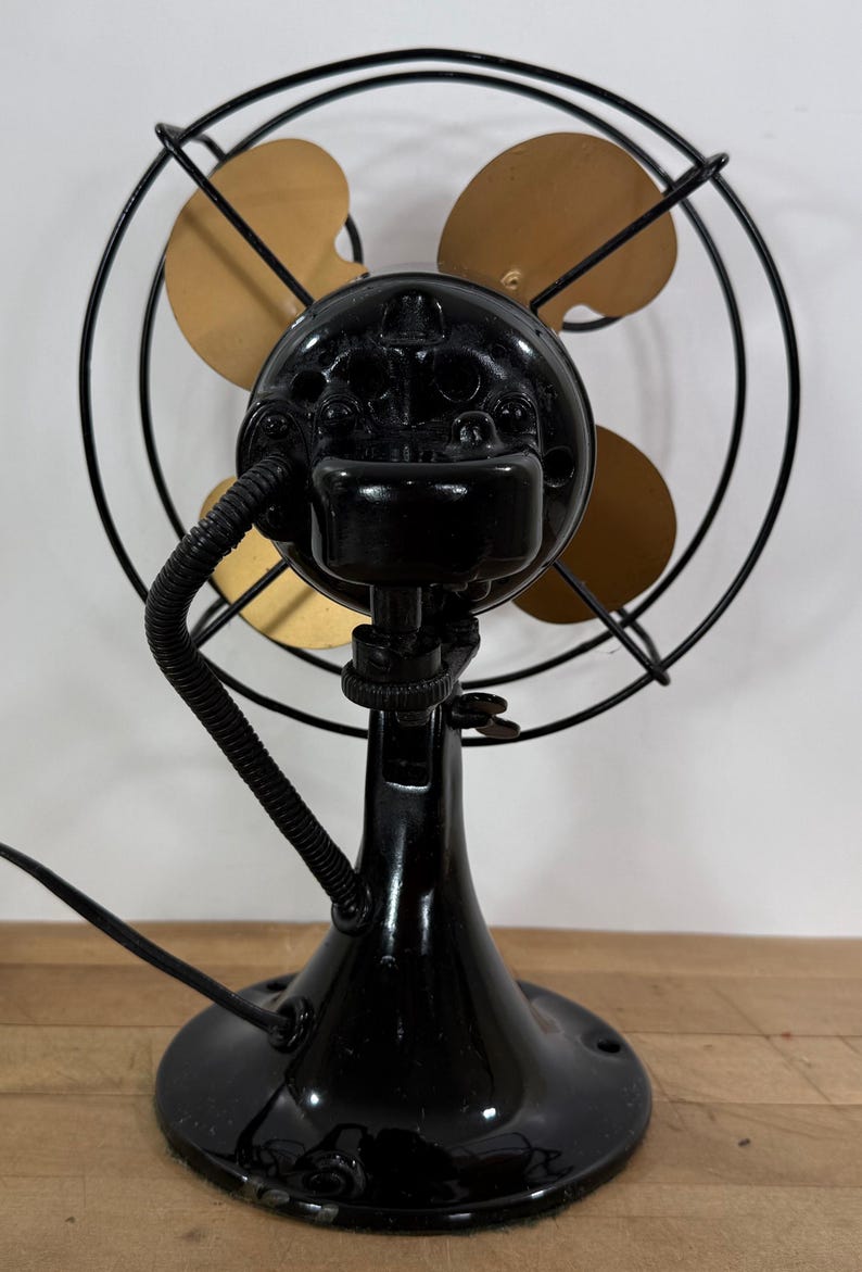 May include: A vintage black electric fan with three gold-coloured blades, enclosed in a black wire cage. The fan has a black base and motor housing, with a flexible cord extending from the back. The fan is a classic design.