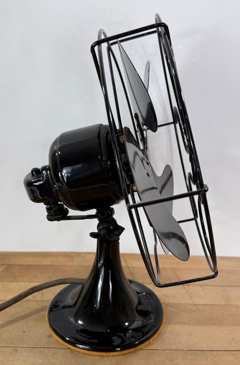 Pu&ograve; includere: Un ventilatore elettrico nero vintage con una gabbia e tre pale. Il ventilatore ha una finitura lucida e una base rotonda. Le pale sono angolate e il ventilatore &egrave; posizionato su una superficie di legno.