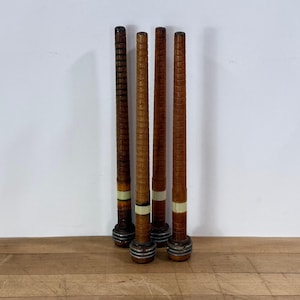 May include: Four antique wooden spools with a cylindrical shape. The spools are various shades of brown, with some featuring decorative bands of white and green. The spools are approximately 10 inches tall and rest on a wooden surface.