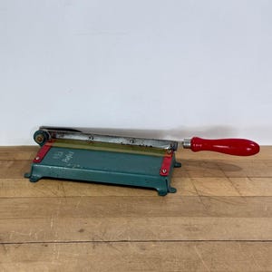May include: A vintage metal paper cutter featuring a teal green base with red accents and a long, sharp blade. The handle is made of smooth, bright red wood. The word "Perfect" is printed in white on the front of the teal metal base.