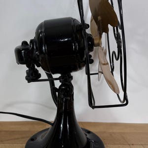 May include: A vintage black electric fan with a round base, a curved stand, and a cage protecting the blades. The fan blades are a light gold colour. The fan is plugged into an electrical outlet.