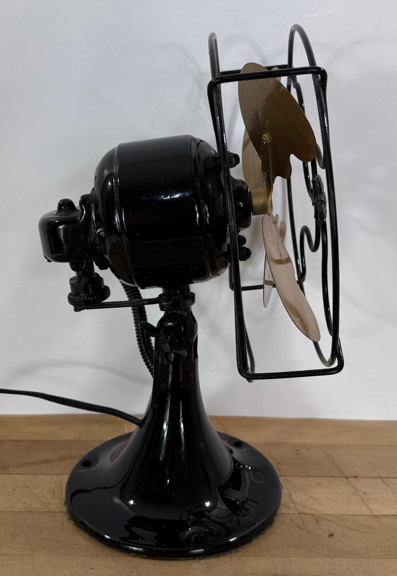 May include: A vintage black electric fan with a round base, a curved stand, and a cage protecting the blades. The fan blades are a light gold colour. The fan is plugged into an electrical outlet.