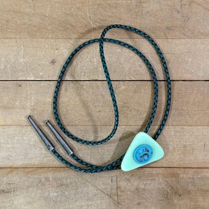 May include: A bolo tie featuring a braided dark teal cord with silver-toned metal tips. The central slide is a triangular light mint green piece with a circular, textured bright blue accent button centered on the front surface of the triangle.