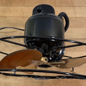 May include: Black electric fan with two bronze-coloured blades, encased in a black wire cage. The fan's motor housing is black, and the power cord is visible. The fan is set against a wooden background.